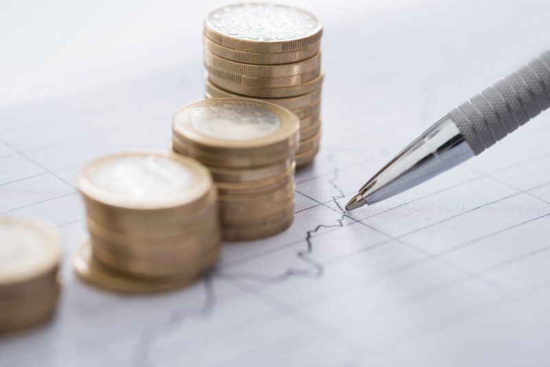 Cropped image of businessman's hand with pen analyzing line and coins graphs on desk